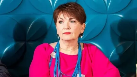 Zorica Kondža u ružičastoj bluzi s plavom ogrlicom ispred plave pozadine