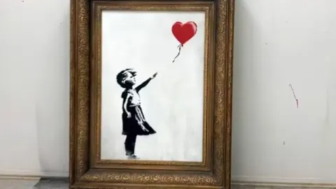 Banksyjeva slika djevojčice s crvenim balonom u zlatnom okviru na bijelom zidu.