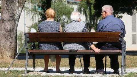 Tri starije osobe sjede na drvenoj klupi u parku okrenute leđima.