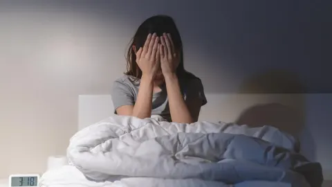 Žena sjedi u krevetu s rukama preko lica u tamnoj sobi.