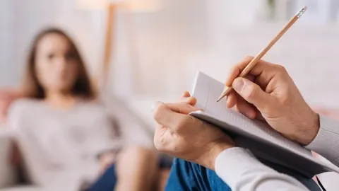 Ruka psihologa piše u bilježnicu dok pacijentica sjedi u pozadini.