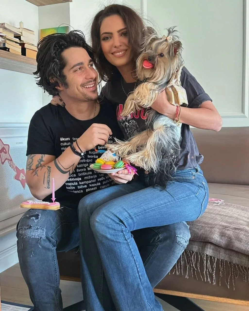 Igor Drvenkar i njegova partnerica sjede na trosjedu uz psa i tanjur šarenih kolačića.