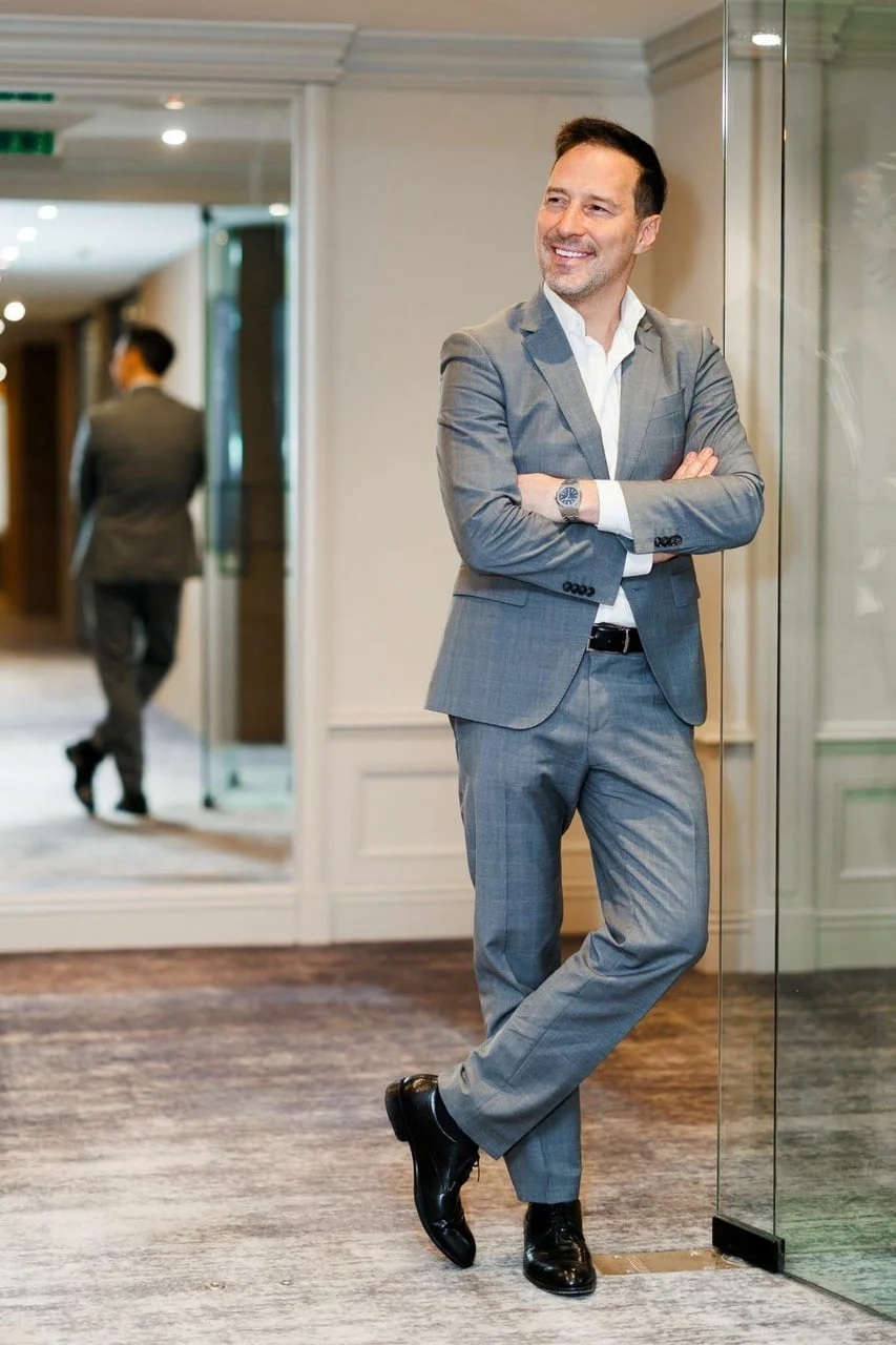 Peđa Jovanović stoji naslonjen na stakleni zid u elegantnom sivom odijelu.