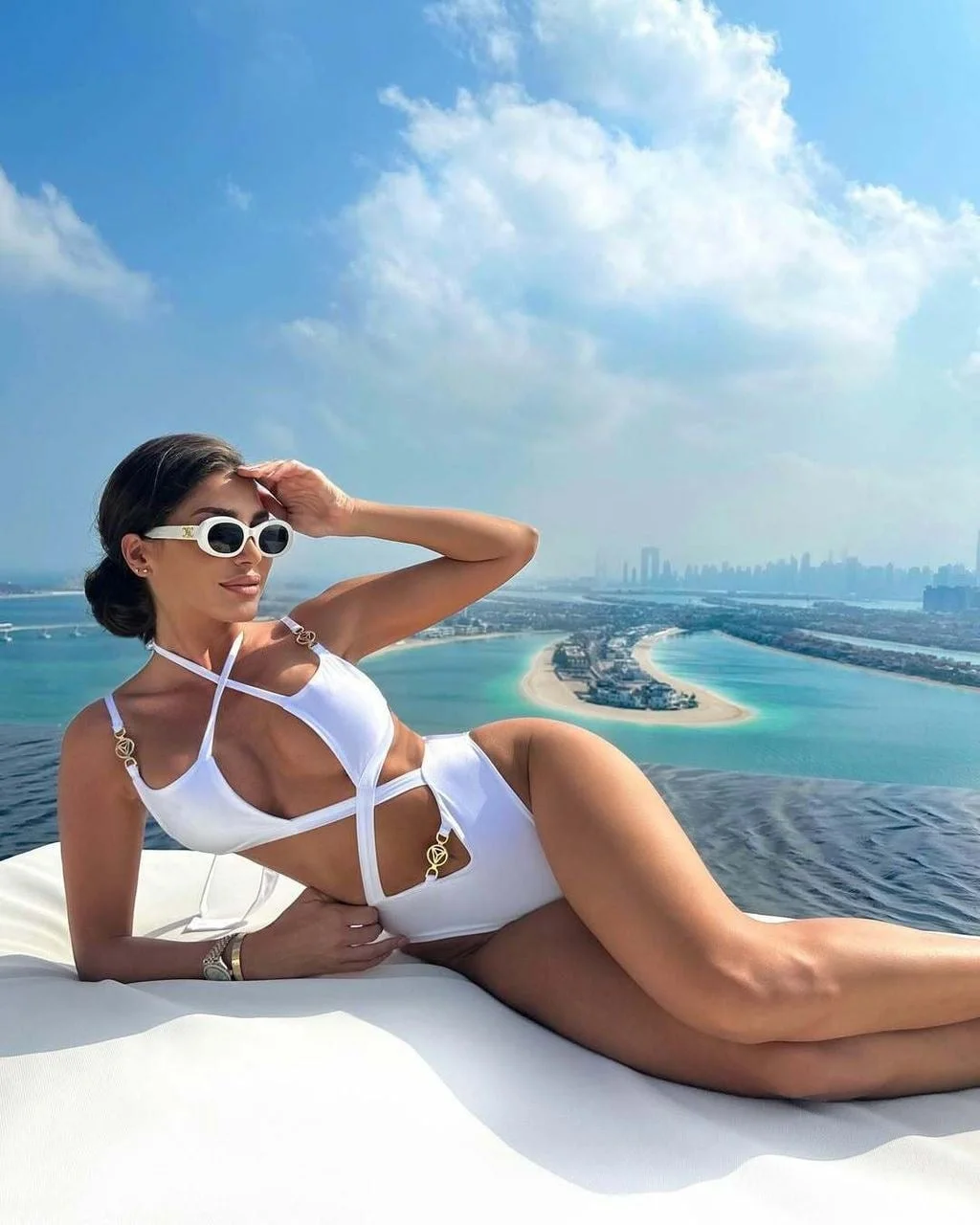 Žena u bijelom kupaćem kostimu pozira na ležaljci uz bazen s pogledom na more.