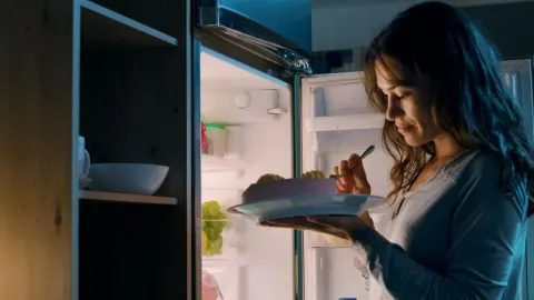 Mlada žena jede tortu ispred otvorenog hladnjaka u tamnoj kuhinji tijekom noći.