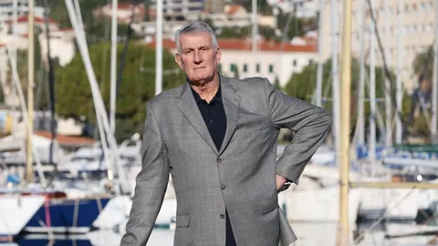 Kрупni plan starijeg muškarca u sivom saku pred marinom s jedrilicama