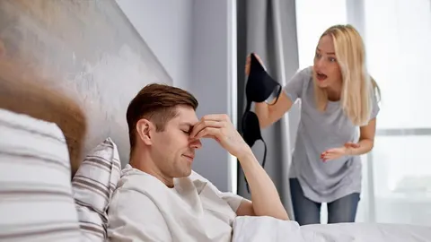 Muškarac u krevetu drži ruku na čelu dok žena u pozadini pokazuje ženski donji ruba