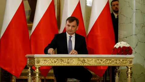 Muškarac u tamnom odelu sedi za ukrašenim stolom ispred poljskih zastava