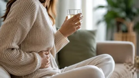 Žena u svijetlom džemperu drži čašu vode i prereće trbuh