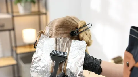 Frizir u crnim rukavicama nanosi boju na plavu kosu pomoću četkice i aluminijskih folija