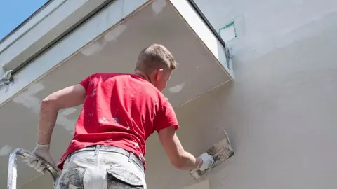 Radnik u crvenoj majici nanosi svijetlo sivi žbuk na vanjski zid kuće