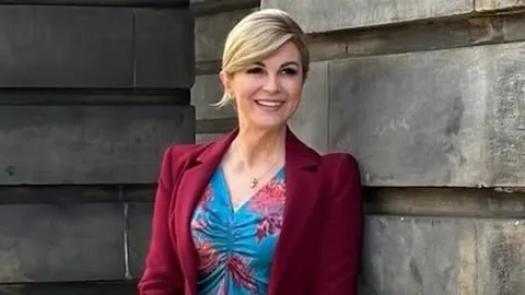 Kolinda Grabar-Kitarović u crvenom saku i plavoj haljini ispred kamenog zida
