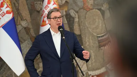 Aleksandar Vučić u tamnoplavom odelu govori pred mikrofonima i zastavama Srbije