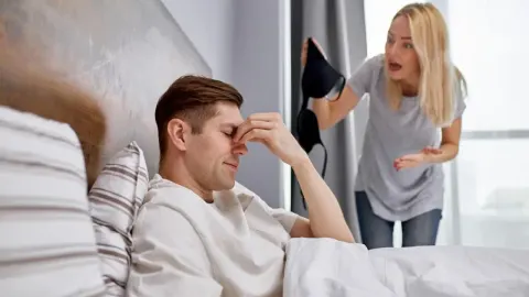 Žena drži komad odjeće i viče na muškarca koji sjedi u krevetu zatvorenih očiju.