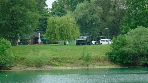 Vojno vozilo i bijeli kombi parkirani na zelenoj površini uz jezero Jarun