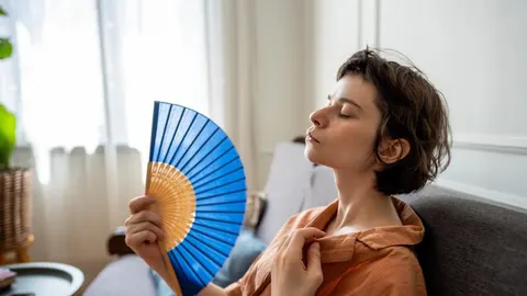 Žena zatvorenih očiju hladi se plavim ručnim lepezom u svijetloj prostoriji