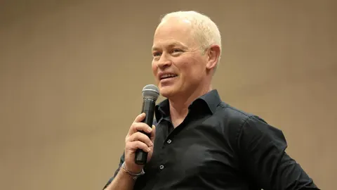 Glumac Neal McDonough u crnoj košulji drži mikrofon i govori