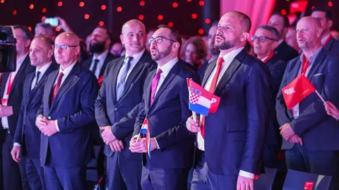 Grupa muškaraca u tamnim odelima na skupu, jedan drži zastavu Hrvatske