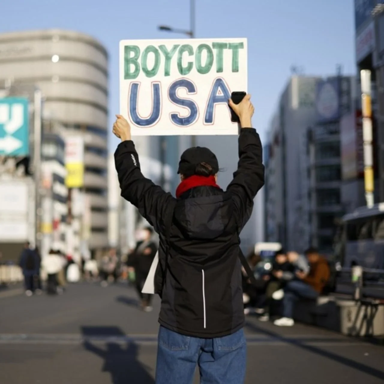 Osoba u crnoj jakni drži natpis 'Boycott USA' na gradskoj ulici