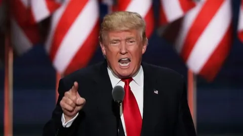 Donald Trump u crnom odelu i crvenoj kravati govori pred američkim zastavama