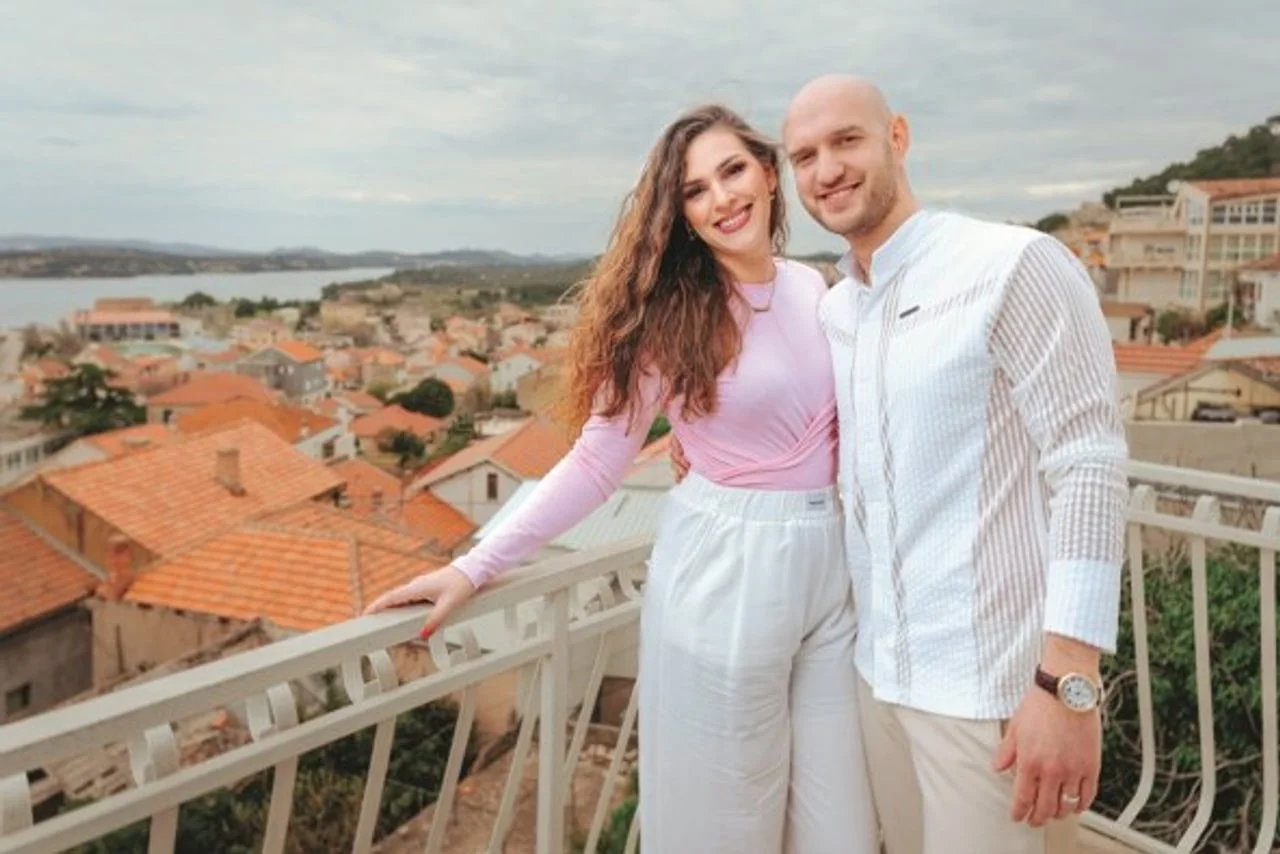 Par u bijeloj odjeći stoji na terasi s pogledom na krovove grada
