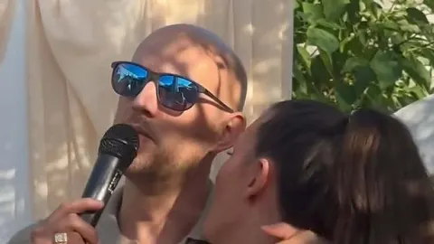 Muškarac u sunčalima pjeva u mikrofon dok ga žena ljubi u obraz