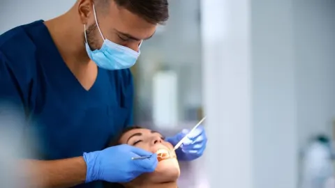 Stomatolog s maskom i rukavicama pregledava usnu šupljinu pacijentice