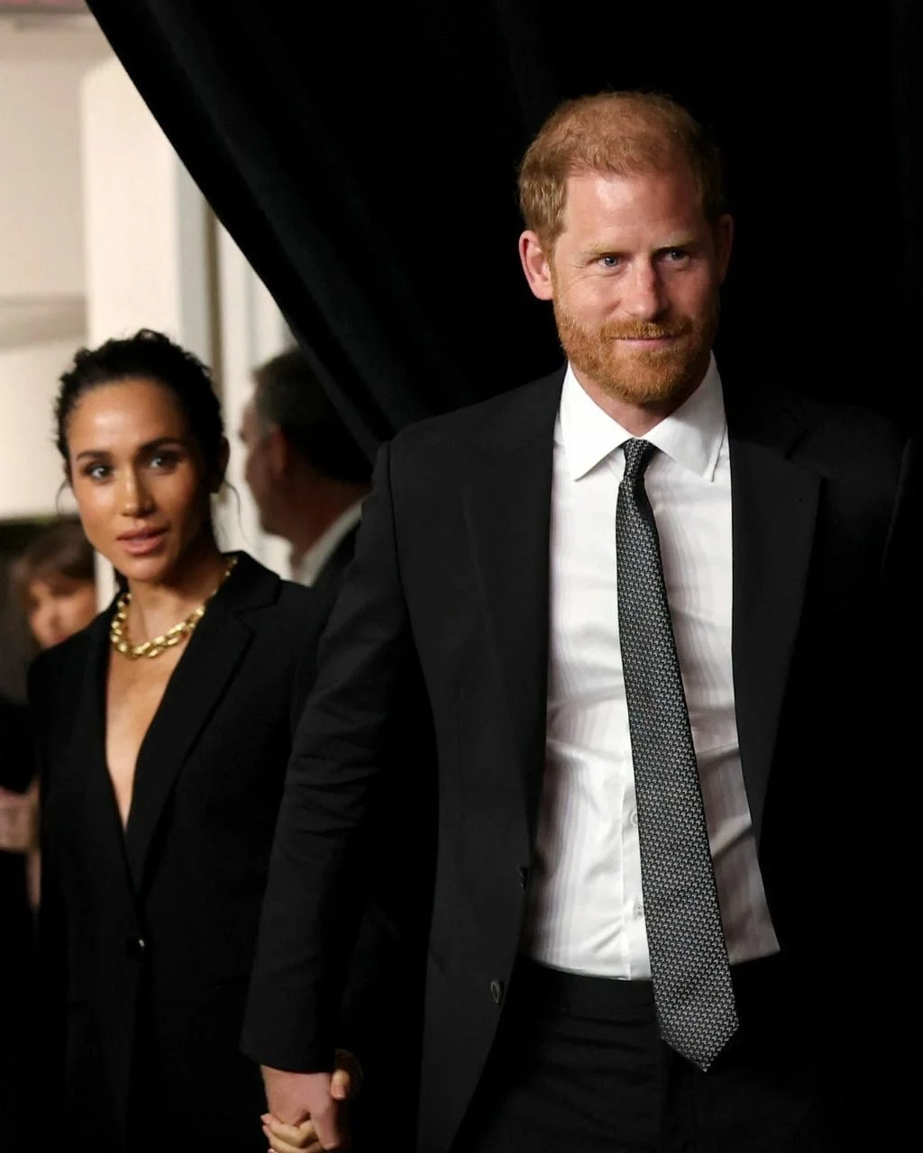 Princ Harry i Meghan Markle drže se za ruke u elegantnim crnim odijelima