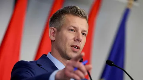 Muškarac u plavom odelu govori za govornicom i pokazuje prstom