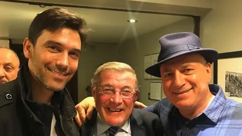 Tri nasmijana muškarca poziraju za zajedničku fotografiju u zatvorenom prostoru