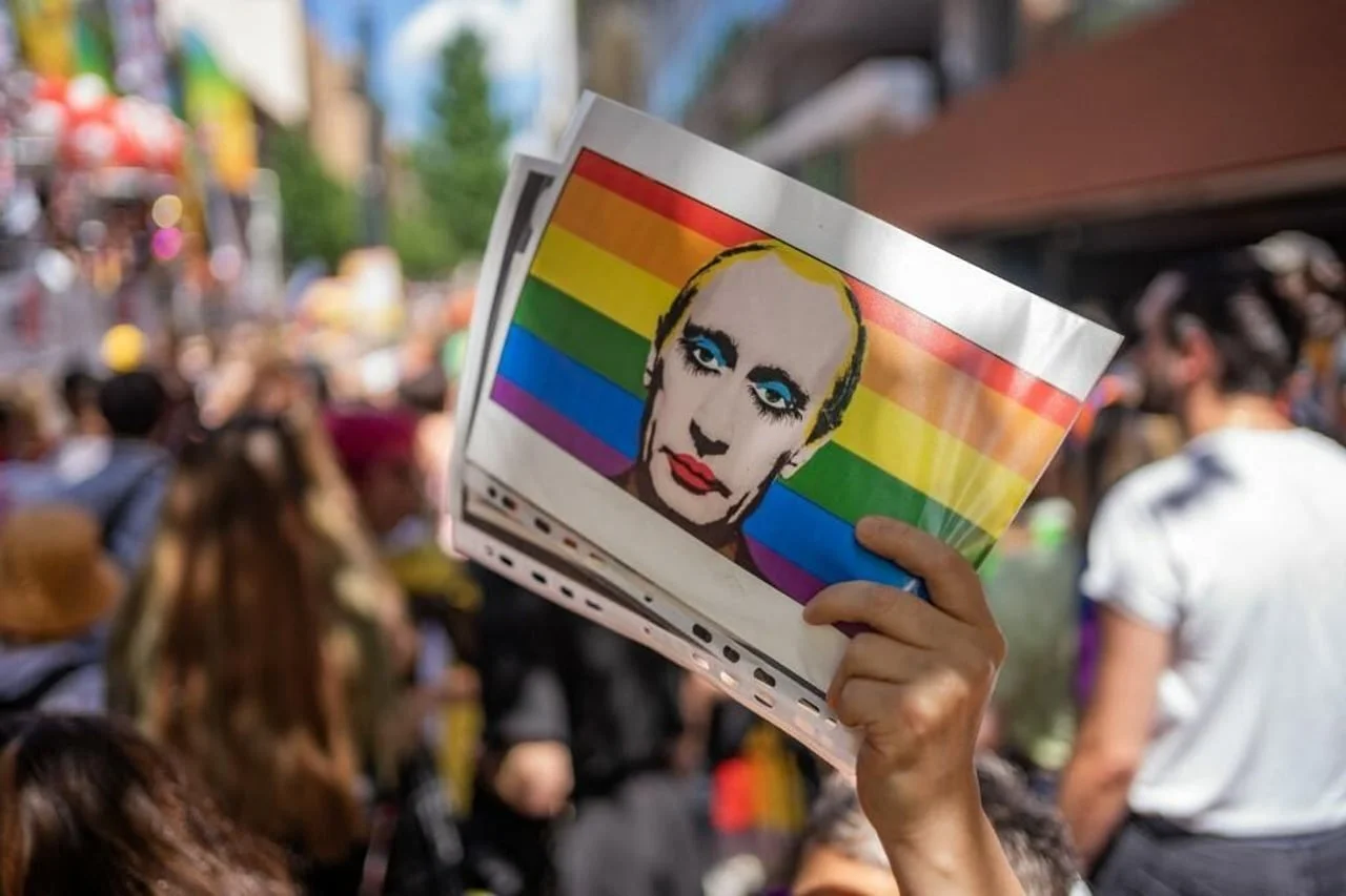 Ruka koja drži satirički plakat Putina s LGBTQ+ zastavom u pozadini