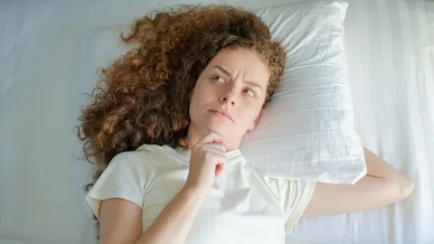 Žena s kovrčavom kosom leži na bijelom jastuku i zamišljeno gleda u stranu