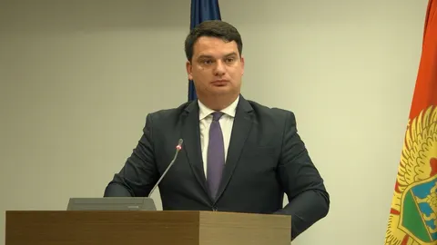 Muškarac u tamnom odjełu i ljubičastoj kravati stoji za govornicom