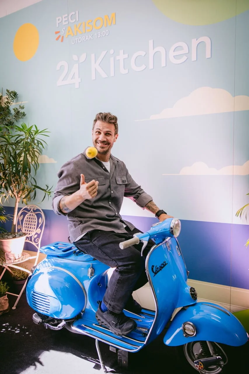 Akis Petretzikis sjedi na plavom Vespa skuteru ispred zida s natpisom