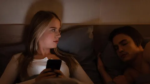 Žena u krevetu tajno gleda u ekran mobitela dok muškarac spava