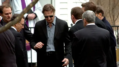 Liam Neeson u crnom odelu i sunčanim naočalama među ljudima