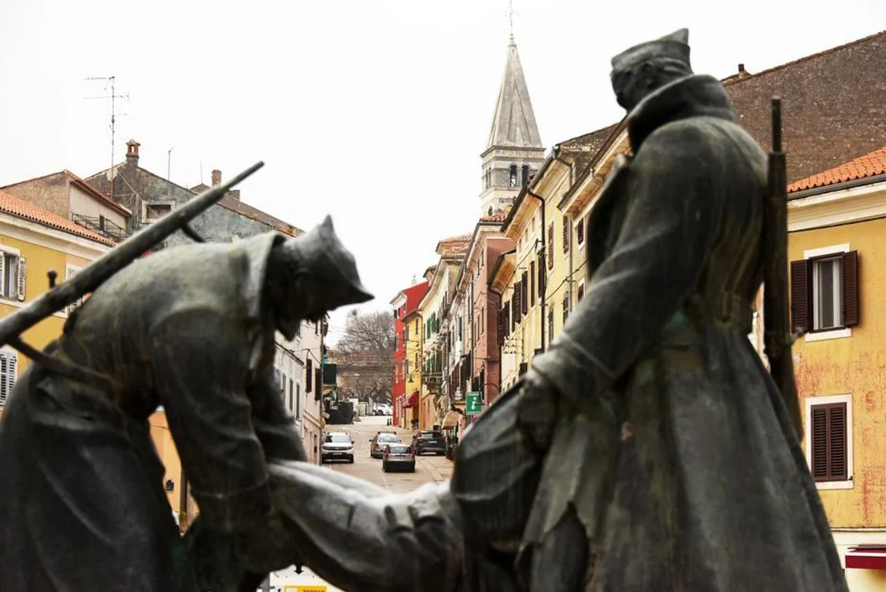 Brončani spomenik u prvom planu s pogledom na ulicu i toranj u pozadini.