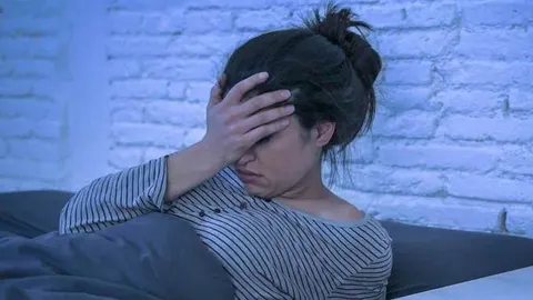 Žena u pstripednoj pidžami leži u krevetu s rukom na čelu u plavičastom svjetlu