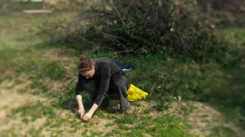 Žena u tamnoj odjeći čuči na travi i radi u vrtu