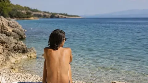 Žena u toplesu sjedi na kamenitoj plaži i gleda u plavo more