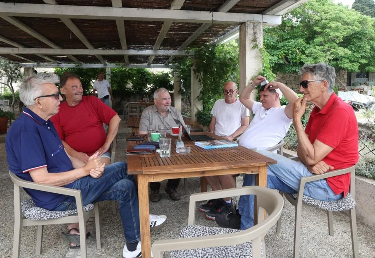 Grupa starijih muškaraca razgovara za drvenim stolom u hladu