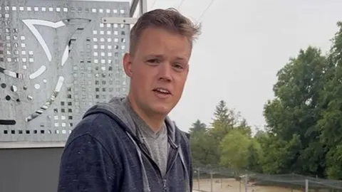 Muškarac u plavoj jakni s kapuljačom stoji ispred metalne skulpture i zelenila