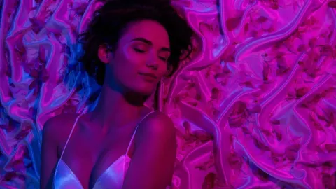 Žena leži na svilenoj posteljini pod ljubičastim i plavim svjetlom