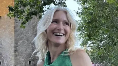 Nasmijana plavokosa žena u zelenoj haljini pred kamenim zidom i drvećem