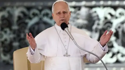 Papa Leo u bijeloj halji govori pred mikrofonom s podignutim rukama.