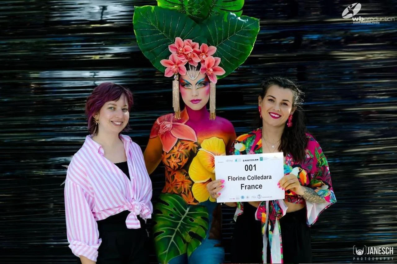 Dvije žene stoje uz model s cvjetnim bodypaintingom i natjecateljskim brojem