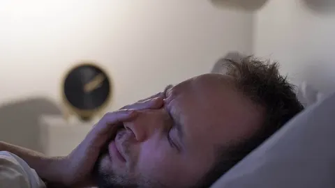 Umoran muškarac s zatvorenim očima leži u krevetu u mračnoj sobi