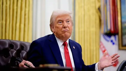 Donald Trump u tamnom odijelu i crvenoj kravati sjedi u uredu
