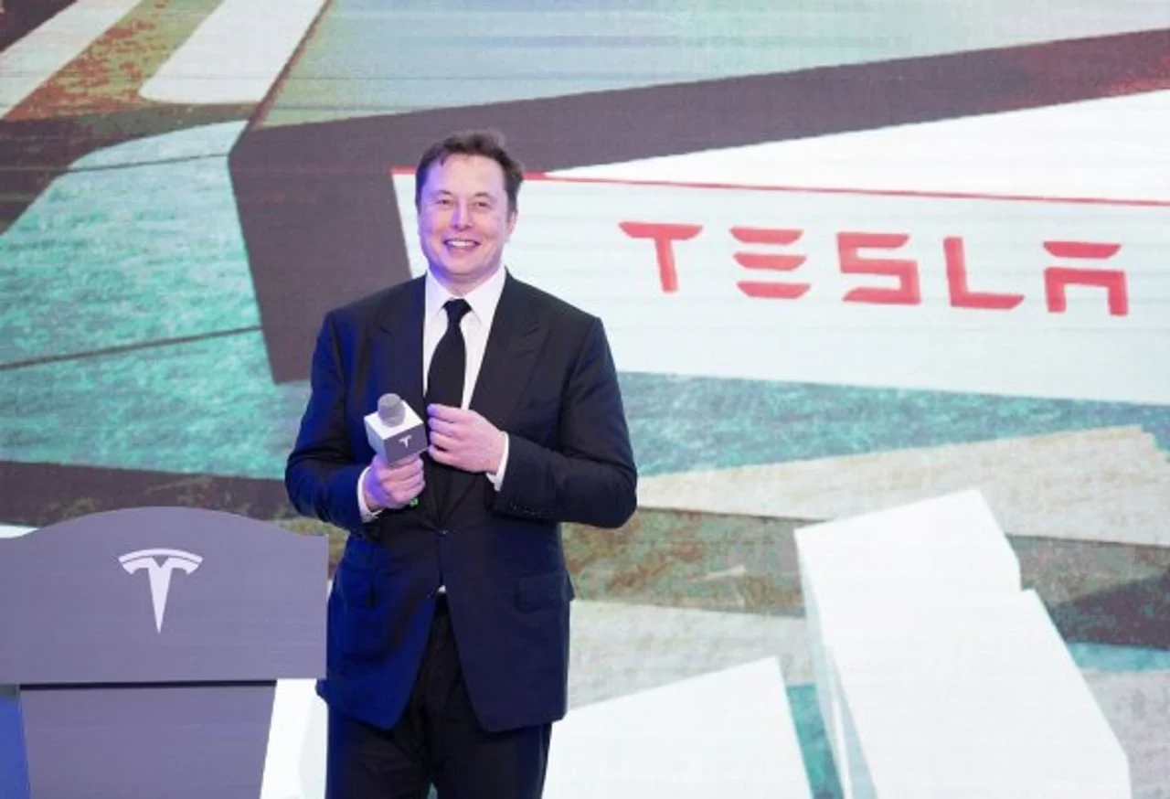 Elon Musk u crnom odelu drži mikrofon ispred Tesla logotipa