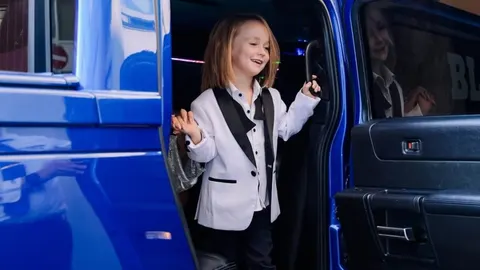 Dječak Bloom u bijelom saku i crnoj mašli izlazi iz plavog automobila